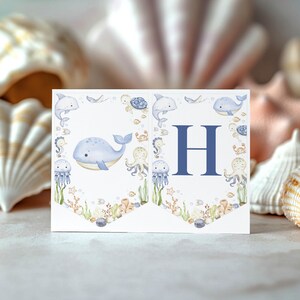 May include: A white banner with a blue letter "H" and watercolor illustrations of marine life, including whales, dolphins, and jellyfish. The banner is decorated with sea creatures and coral, perfect for a nautical-themed event.