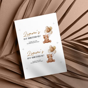 May include: A white rectangular card featuring two identical prints of a brown teddy bear holding a cluster of balloons with the number 1 on the centre balloon. The text reads Liam's 1ST BIRTHDAY! THANK YOU FOR CELEBRATING WITH ME! in brown and black.