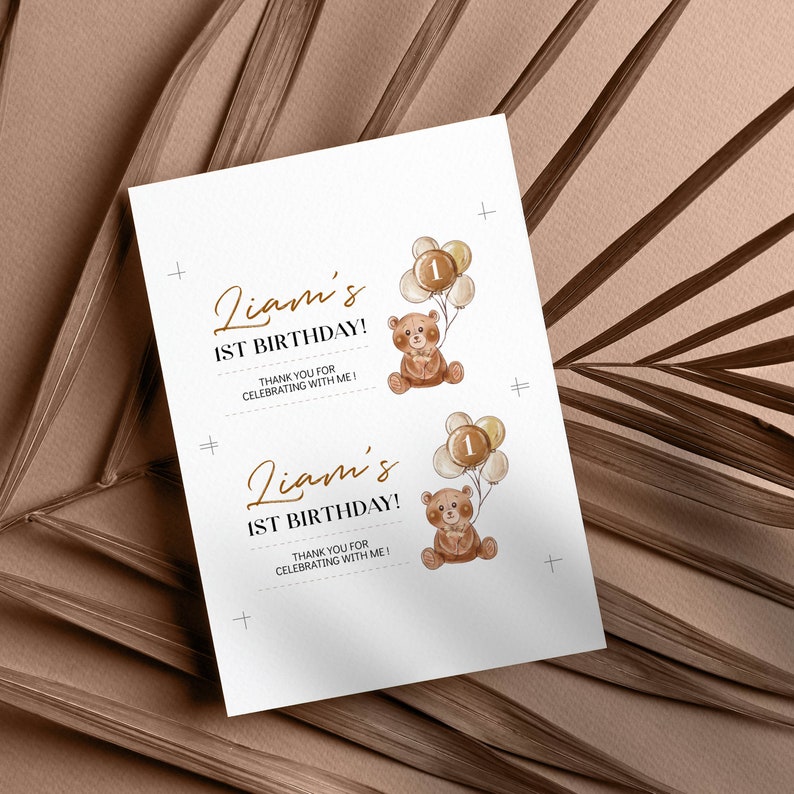 May include: A white rectangular card featuring two identical prints of a brown teddy bear holding a cluster of balloons with the number 1 on the centre balloon. The text reads Liam's 1ST BIRTHDAY! THANK YOU FOR CELEBRATING WITH ME! in brown and black.