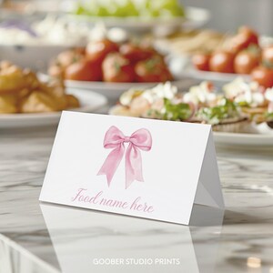 May include: A white folded card featuring a watercolor illustration of a light pink bow centered at the top. Below the bow, the text "Food name here" is printed in a light pink cursive script. The card is displayed in a tent-fold style.