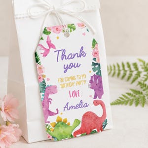 K&ouml;nnte beinhalten: Ein wei&szlig;es, rechteckiges Geschenkanh&auml;nger-Etikett mit abgerundeten oberen Ecken, verziert mit Aquarellillustrationen von rosa, gr&uuml;nen und violetten Dinosauriern, rosa Blumen und gr&uuml;nen Bl&auml;ttern. Der Text lautet: Thank you FOR COMING TO MY BIRTHDAY PARTY! LOVE, Amelia.