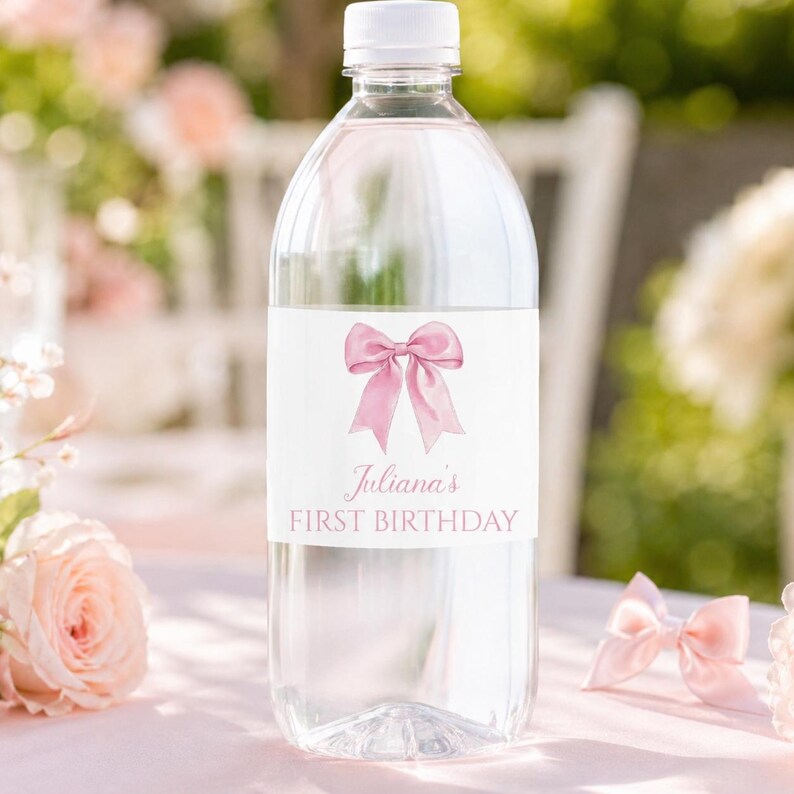 Puede incluir: Una botella de agua de pl&aacute;stico transparente con una etiqueta blanca que presenta una ilustraci&oacute;n de un lazo rosa. El texto en la etiqueta dice Juliana's FIRST BIRTHDAY en una fuente rosa cursiva y de bloque. La botella tiene una tapa blanca.