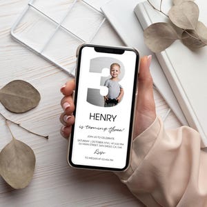 Op de afbeelding: Een smartphone met een digitale verjaardagsuitnodiging met een groot cijfer drie waarin een foto van een jong kind is verwerkt. De tekst luidt Henry is turning three, join us to celebrate, Saturday October 15th at 2:00 PM, 123 Main Street, San Diego, CA, 1345, RSVP to Megan at 123.456.789.