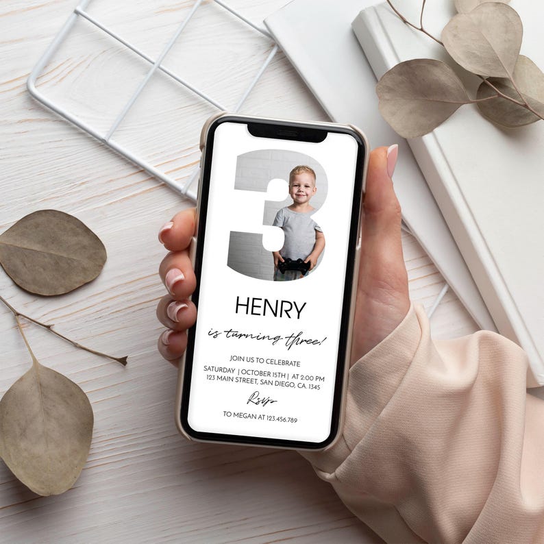 Op de afbeelding: Een smartphone met een digitale verjaardagsuitnodiging met een groot cijfer drie waarin een foto van een jong kind is verwerkt. De tekst luidt Henry is turning three, join us to celebrate, Saturday October 15th at 2:00 PM, 123 Main Street, San Diego, CA, 1345, RSVP to Megan at 123.456.789.
