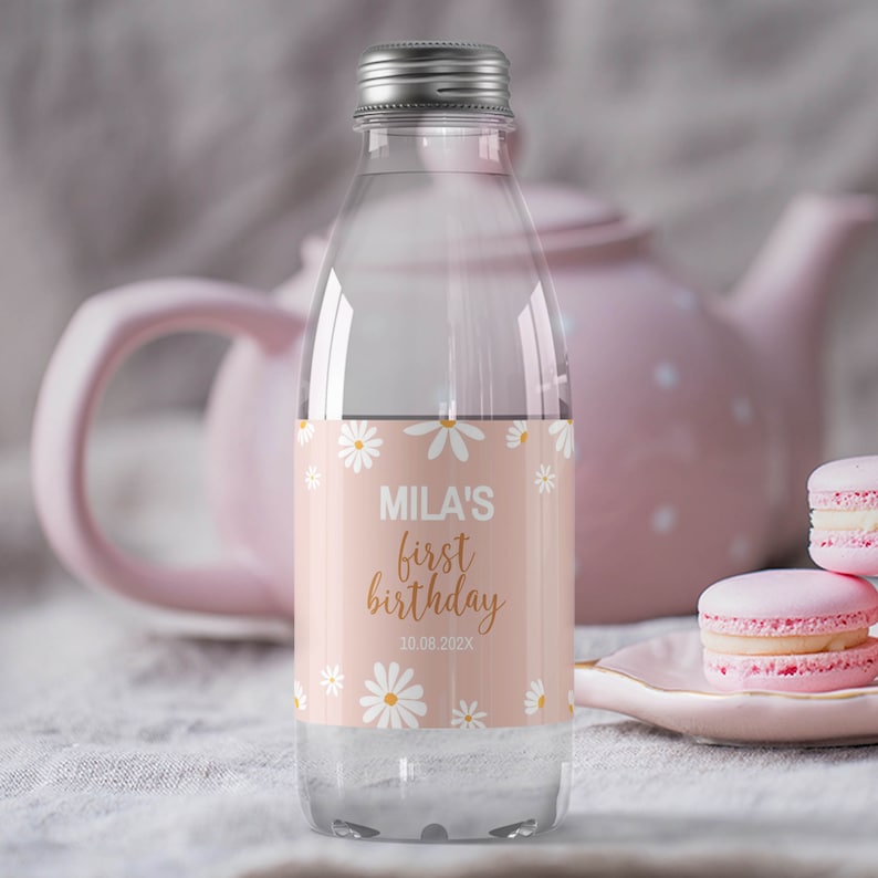 May include: A clear plastic water bottle with a silver screw cap, featuring a light pink label decorated with white daisy flowers. The label displays the text "MILA'S first birthday" in white and gold script, along with the date "10.08.202X".