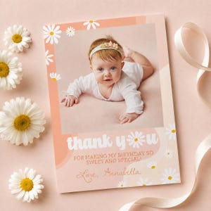 May include: A rectangular thank you card with a peach-colored border featuring white daisy illustrations. The center displays a photograph of a baby wearing a white long-sleeved outfit and a floral headband. Text reads: thank you FOR MAKING MY BIRTHDAY SO SWEET AND SPECIAL! Love, Annabella.