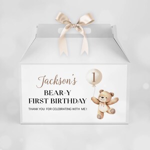 May include: A white gable box with a beige satin ribbon handle. The front features a brown teddy bear illustration holding a balloon with the number 1, and text reading: Jackson's BEAR-Y FIRST BIRTHDAY THANK YOU FOR CELEBRATING WITH ME!