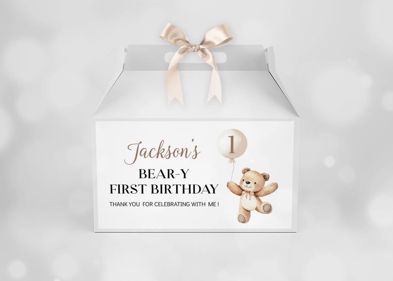 May include: A white gable box with a beige satin ribbon handle. The front features a brown teddy bear illustration holding a balloon with the number 1, and text reading: Jackson's BEAR-Y FIRST BIRTHDAY THANK YOU FOR CELEBRATING WITH ME!