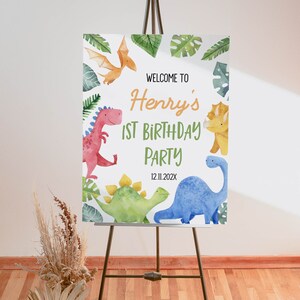 Op de afbeelding: Een rechthoekig welkomstbord voor een verjaardagsfeest met aquarelillustraties van diverse dinosaurussen en tropische bladeren. De tekst luidt: WELCOME TO Henry's 1ST BIRTHDAY PARTY 12.11.202X. Het bord staat op een zwarte metalen schildersezel.