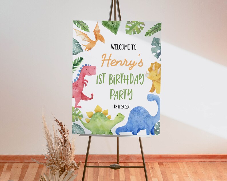 Op de afbeelding: Een rechthoekig welkomstbord voor een verjaardagsfeest met aquarelillustraties van diverse dinosaurussen en tropische bladeren. De tekst luidt: WELCOME TO Henry's 1ST BIRTHDAY PARTY 12.11.202X. Het bord staat op een zwarte metalen schildersezel.