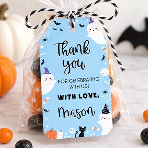 Pu&ograve; includere: Un'etichetta regalo rettangolare azzurra legata con uno spago bianco e nero a un sacchetto di plastica trasparente pieno di caramelle. L'etichetta presenta illustrazioni di fantasmi bianchi con cappelli viola e neri, gatti neri, zucche e il testo Thank you for celebrating with us! With love, Mason.