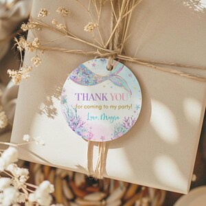 May include: Round thank you tag with a mermaid tail and the text "THANK YOU for coming to my party! Love, Mayra" in pink, purple, and blue lettering on a light blue background with gold glitter.