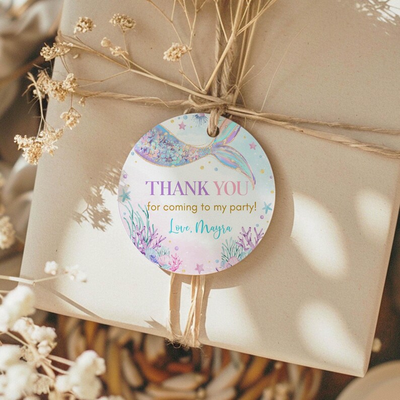 May include: Round thank you tag with a mermaid tail and the text "THANK YOU for coming to my party! Love, Mayra" in pink, purple, and blue lettering on a light blue background with gold glitter.