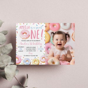 May include: A pink and white donut-themed first birthday party invitation with a photo of a baby girl surrounded by donuts. The invitation says "Amelia is a sweet one!" and includes the date, time, and location of the party.