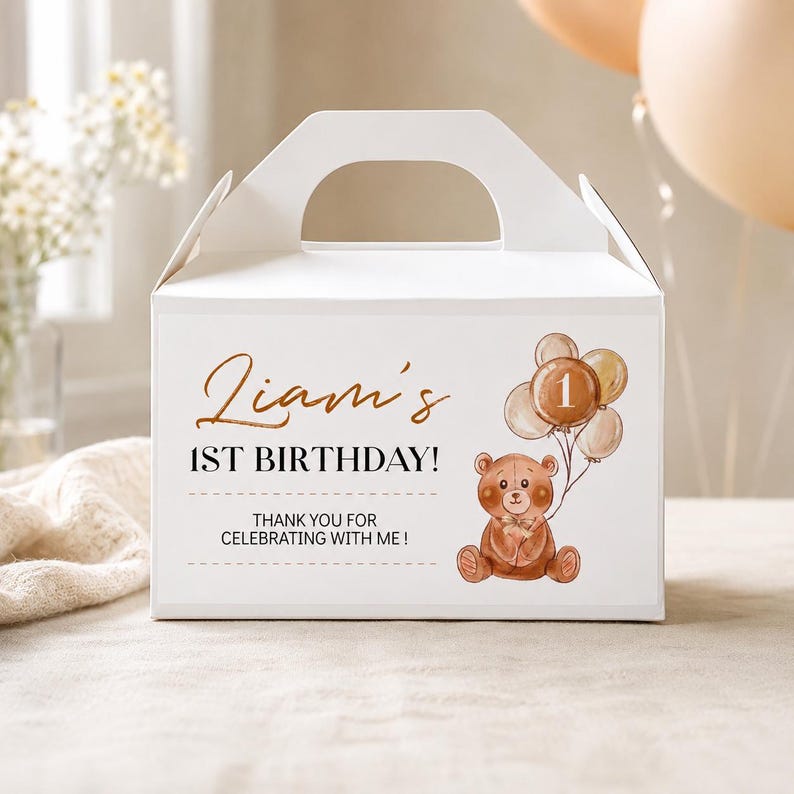 May include: A white cardboard gable box featuring a brown teddy bear illustration holding a cluster of balloons with the number 1. The text reads: Liam's 1ST BIRTHDAY! THANK YOU FOR CELEBRATING WITH ME! The design uses brown and black lettering.