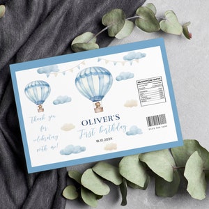 May include: A light blue birthday party favor with watercolor hot air balloons, clouds, and text. The text reads "Thank you for celebrating with me!" and "Oliver's First Birthday 18.10.202X". Includes a nutrition facts label.