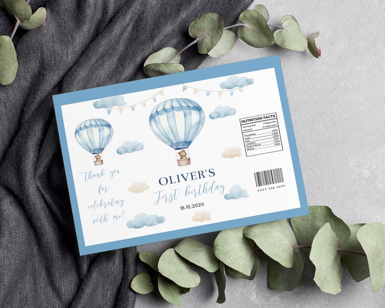 May include: A light blue birthday party favor with watercolor hot air balloons, clouds, and text. The text reads "Thank you for celebrating with me!" and "Oliver's First Birthday 18.10.202X". Includes a nutrition facts label.