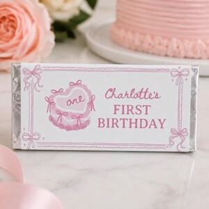 May include: A rectangular candy bar wrapper featuring a white background with pink decorative borders and bows. The design includes a pink illustration of a heart-shaped cake with the word one and the text Charlotte's FIRST BIRTHDAY in pink.