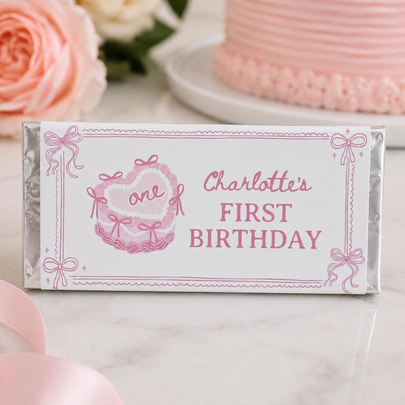 May include: A rectangular candy bar wrapper featuring a white background with pink decorative borders and bows. The design includes a pink illustration of a heart-shaped cake with the word one and the text Charlotte's FIRST BIRTHDAY in pink.