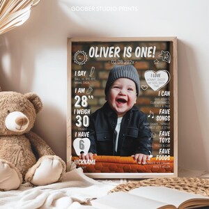 May include: A framed milestone poster featuring a photograph of a smiling baby wearing a gray beanie and black shirt. The text reads OLIVER IS ONE, 02.08.202x, 28 inches, 30 pounds, 6 teeth, and lists favorite songs, toys, foods, and nicknames.