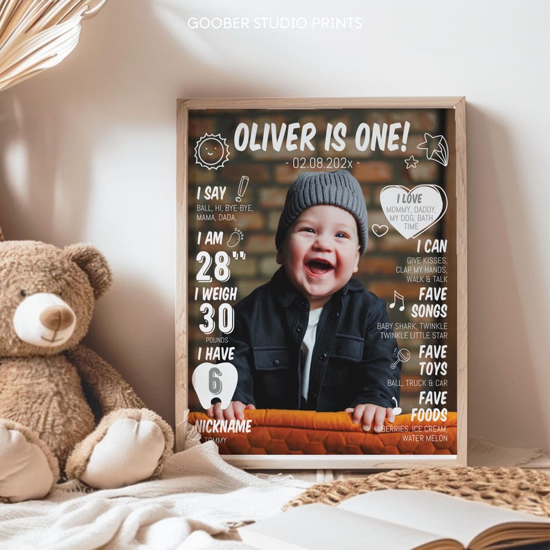 May include: A framed milestone poster featuring a photograph of a smiling baby wearing a gray beanie and black shirt. The text reads OLIVER IS ONE, 02.08.202x, 28 inches, 30 pounds, 6 teeth, and lists favorite songs, toys, foods, and nicknames.