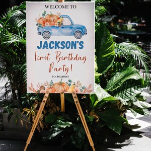 May include: A rectangular white sign on a wooden easel featuring a blue truck illustration carrying pumpkins, with the text: WELCOME TO, one, JACKSON'S, First Birthday Party!, and 10.10.202X, decorated with watercolour pumpkins and autumn foliage.