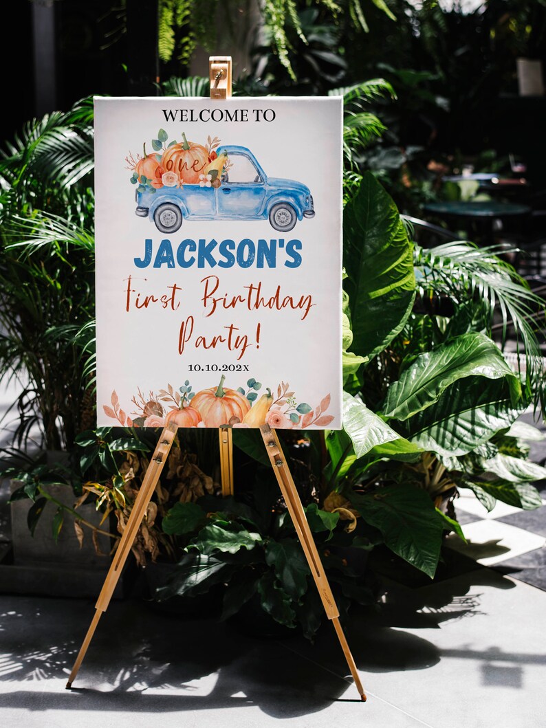 May include: A rectangular white sign on a wooden easel featuring a blue truck illustration carrying pumpkins, with the text: WELCOME TO, one, JACKSON'S, First Birthday Party!, and 10.10.202X, decorated with watercolour pumpkins and autumn foliage.