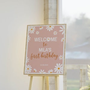 Op de afbeelding: Een rechthoekig bord met een oudroze achtergrond en witte madeliefjesillustraties. De tekst luidt: WELCOME to MILA'S first birthday 10.08.202X. Het bord staat op een lichtgekleurde houten driepoot-ezelstandaard voor evenementenbordjes.