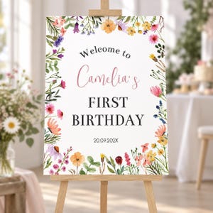 K&ouml;nnte beinhalten: Ein rechteckiges wei&szlig;es Schild auf einer Holzstaffelei mit einem Aquarell-Blumenrand aus rosa, orangefarbenen, gelben und violetten Bl&uuml;ten. Der Text lautet Welcome to Camelia's FIRST BIRTHDAY 20.09.202X in schwarzen und rosa Schriftarten.
