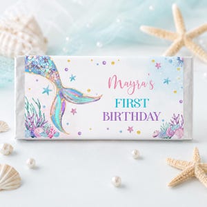 Op de afbeelding: Een rechthoekige wikkel voor een chocoladereep met een aquarel zeemeermin staart in blauwe, paarse en gouden tinten. De tekst luidt Mayra's FIRST BIRTHDAY in roze en turquoise letters, omringd door illustraties van koraal en zeesterren.