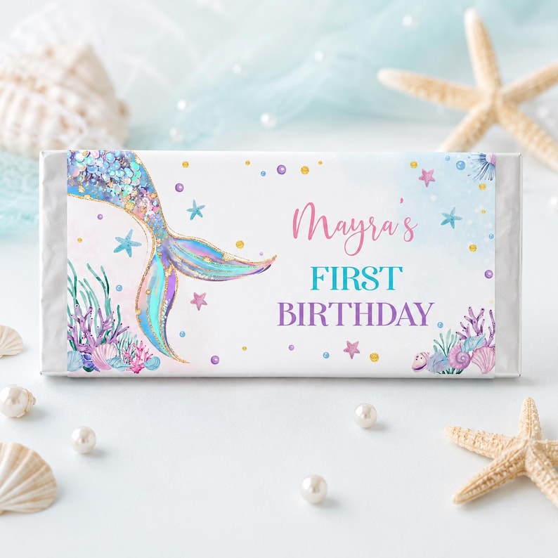 Op de afbeelding: Een rechthoekige wikkel voor een chocoladereep met een aquarel zeemeermin staart in blauwe, paarse en gouden tinten. De tekst luidt Mayra's FIRST BIRTHDAY in roze en turquoise letters, omringd door illustraties van koraal en zeesterren.