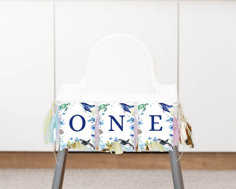 May include: A high chair banner featuring three white panels with the blue letters O, N, and E. The design includes watercolor illustrations of whales, sea turtles, fish, and coral reefs, accented with colorful fabric tassels on the sides.