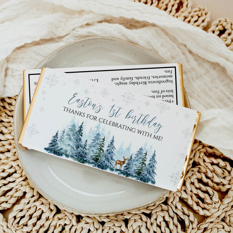 May include: A rectangular chocolate bar wrapper featuring a watercolor forest of blue and green pine trees, a small deer, and snowflakes. The text reads: Easton's 1st birthday, THANKS FOR CELEBRATING WITH ME! The back lists ingredients like birthday magic.