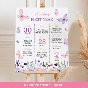 May include: A white milestone poster titled Amelia's First Year, featuring pink and purple butterflies and floral illustrations. The text includes weight in pounds, height in inches, favorite foods, songs, toys, and milestones for a one-year-old.
