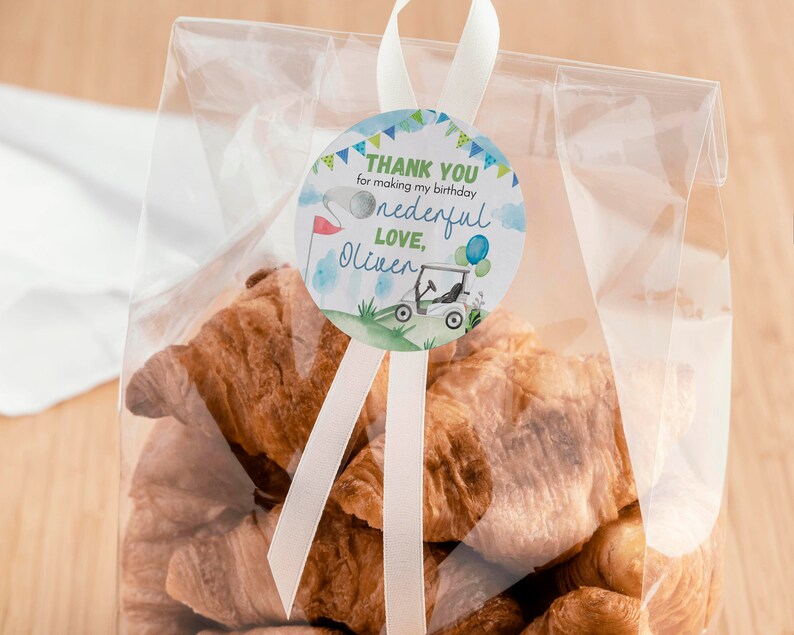 May include: A clear plastic bag filled with golden-brown croissants, sealed with a white ribbon and a round sticker. The sticker features a golf cart, a flag, and the text: THANK YOU for making my birthday onederful LOVE, Oliver.