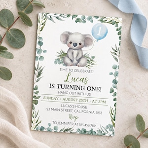 May include: A rectangular birthday invitation card featuring a watercolor illustration of a koala holding a light blue balloon with the number one. The card is framed by green eucalyptus leaves and includes text details for Lucas's first birthday party.