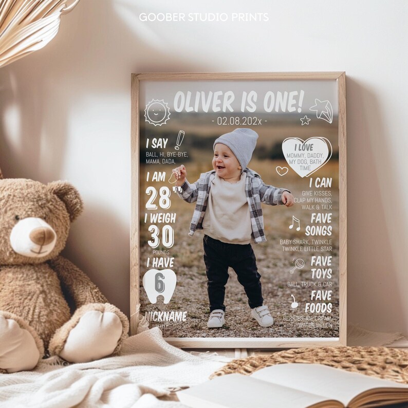 May include: A rectangular framed milestone poster featuring a photograph of a toddler wearing a gray beanie, plaid shirt, and dark pants. The white text includes 'OLIVER IS ONE!', 'I SAY', 'I AM 28"', 'I WEIGH 30 POUNDS', 'I HAVE 6 TEETH', and 'I LOVE'.