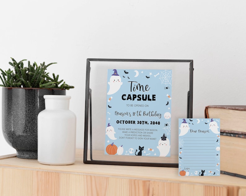 May include: A light blue time capsule sign and matching note card featuring Halloween illustrations of ghosts in hats, pumpkins, a black cat, and spiderwebs. The text reads Time Capsule to be opened on Mason's 18th birthday October 30th, 2040.
