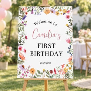 K&ouml;nnte beinhalten: Ein rechteckiges Willkommensschild mit einem aquarellierten Blumenrand aus verschiedenen bunten Wildblumen. Der Text lautet Welcome to Camelia's FIRST BIRTHDAY 20.09.202X. Das Schild steht auf einer h&ouml;lzernen Staffelei vor einem unscharfen Hintergrund.