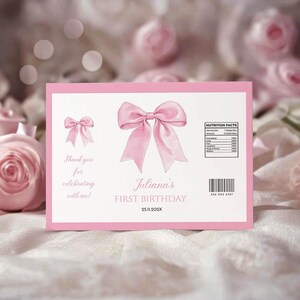 May include: A rectangular pink and white birthday label featuring a large pink bow in the center, a smaller bow on the left, and text reading Juliana's FIRST BIRTHDAY 25.11.202X. It includes a nutrition facts box, a barcode, and thank you text.