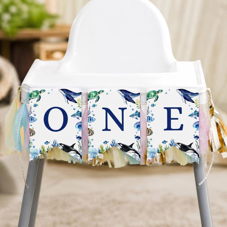 May include: A high chair banner featuring three white pennants with the dark blue letters O, N, and E. Each pennant displays watercolor illustrations of sea creatures including whales, sea turtles, and fish, accented with pastel fringe tassels.