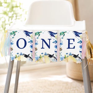 May include: A high chair banner featuring three white rectangular panels with the dark blue letters O, N, and E. The panels are decorated with watercolor illustrations of sea creatures including whales, turtles, and fish, finished with colorful tassels.