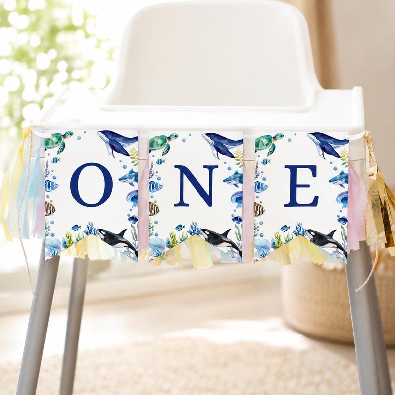May include: A high chair banner featuring three white rectangular panels with the dark blue letters O, N, and E. The panels are decorated with watercolor illustrations of sea creatures including whales, turtles, and fish, finished with colorful tassels.
