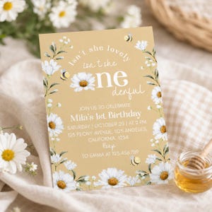 May include: A rectangular birthday invitation card on a gold background featuring white daisy illustrations, green leaves, and small bees. The text reads: Isn't she lovely, isn't she one-derful, Join us to celebrate Mila's 1st Birthday, October 20 at 2 PM.