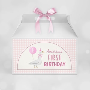 May include: A white gable box with a pink satin ribbon bow on top. The front features a light pink gingham border and a central graphic of a goose wearing a party hat and holding a pink balloon with the number 1, plus the text Amelia's FIRST BIRTHDAY.