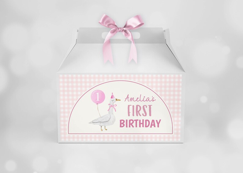 May include: A white gable box with a pink satin ribbon bow on top. The front features a light pink gingham border and a central graphic of a goose wearing a party hat and holding a pink balloon with the number 1, plus the text Amelia's FIRST BIRTHDAY.
