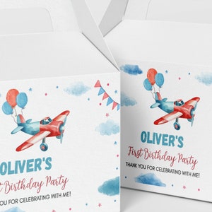 May include: Two white cardboard favor boxes featuring a watercolor illustration of a red and blue airplane with balloons, clouds, and bunting. The text reads: OLIVER'S First Birthday Party THANK YOU FOR CELEBRATING WITH ME! in blue and red ink.