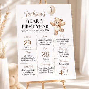 May include: A white milestone birthday sign for Jackson's Bear-y First Year featuring a brown teddy bear illustration, a balloon with the number 1, and text detailing weight in pounds, height in inches, favorite foods, songs, toys, and milestones.