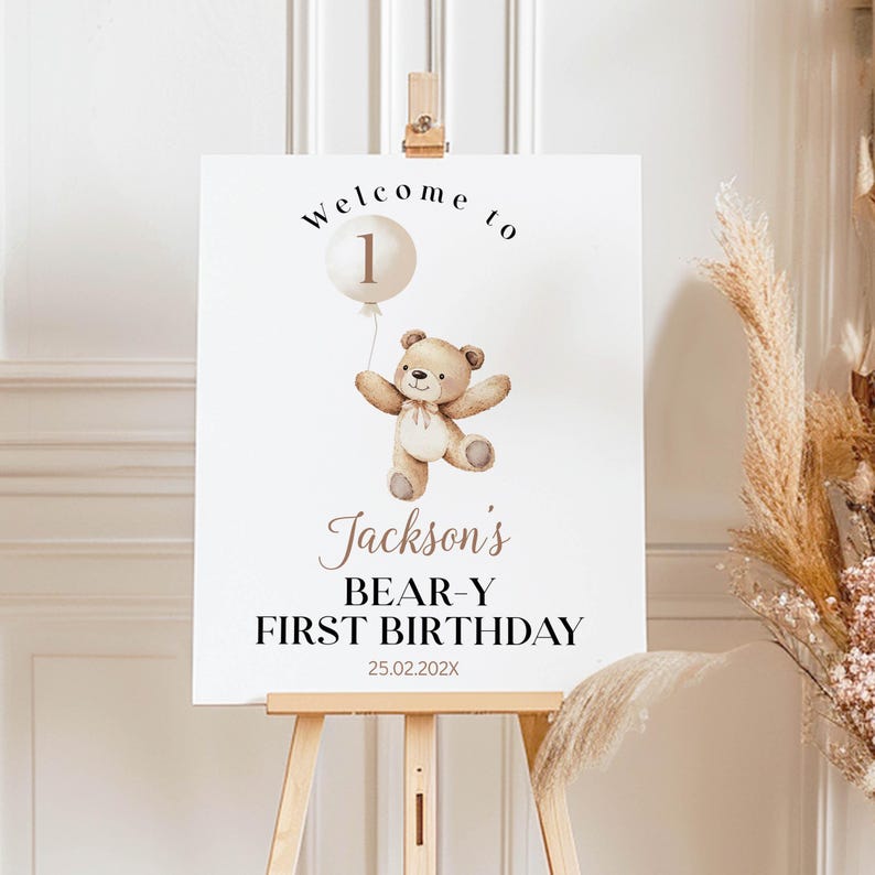May include: A white rectangular welcome sign featuring a brown teddy bear holding a beige balloon with the number 1. The text reads Welcome to Jackson's BEAR-Y FIRST BIRTHDAY 25.02.202X. The sign is displayed on a light wooden easel stand.