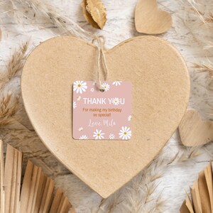 May include: A square pink gift tag with white daisy illustrations and the text "THANK YOU For making my birthday so special! Love, Mila" printed in white and orange. The tag is attached with a jute string to a large, heart-shaped cardboard box.